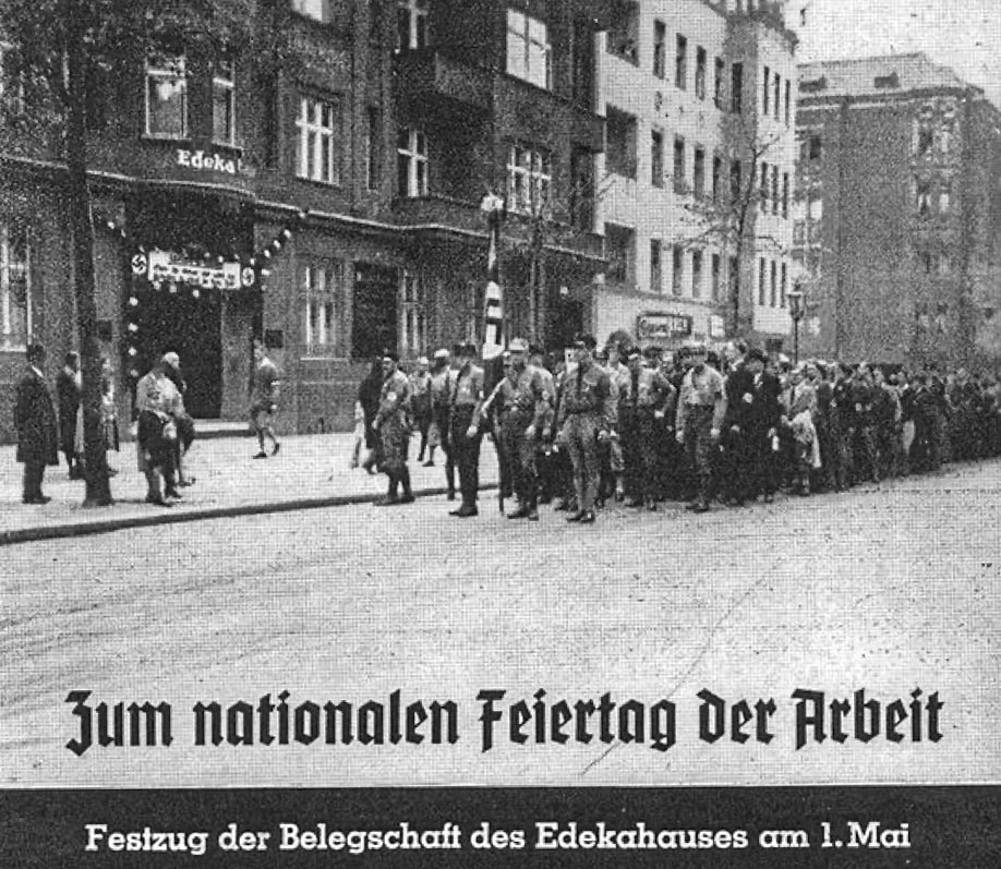 Belegschaft vor der Edeka-Zentrale am 1. Mai 1937. Ein Teil des Aufmarsches trägt Uniformen. An der Edeka-Zentrale sind Hakenkreuz-Symbole zu sehen. In Fraktura ist zu lesen: "Jum nationalen Feiertag Der Arbeit"